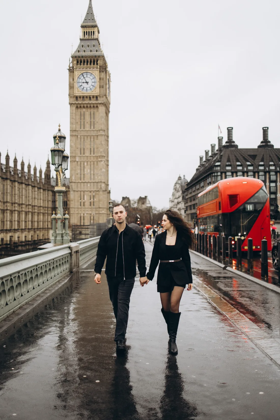 Couple walking hand in hand across wet Westminster Bridge with a red bus passing